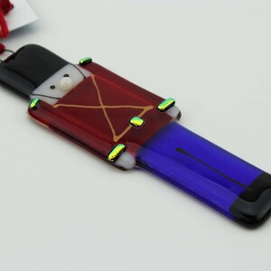 May include: A glass Christmas ornament shaped like a toy soldier. The soldier is wearing a red coat with gold trim, a black hat, and blue pants. The ornament has a white face with a pearl button for a nose.