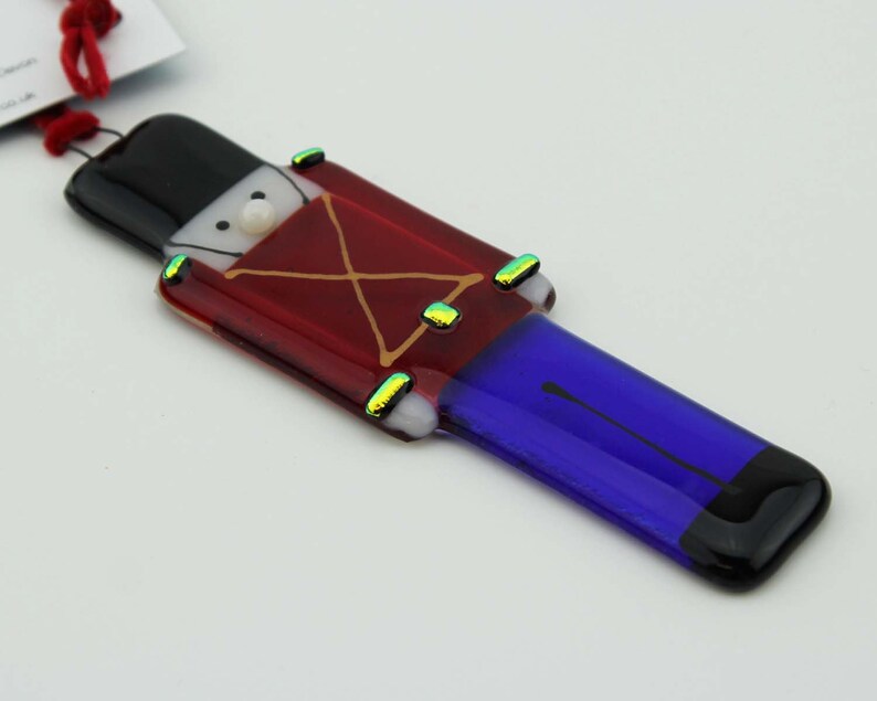 May include: A glass Christmas ornament shaped like a toy soldier. The soldier is wearing a red coat with gold trim, a black hat, and blue pants. The ornament has a white face with a pearl button for a nose.