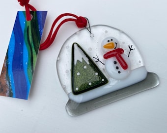 May include: A fused glass ornament shaped like a snow globe featuring a white snowman with a red scarf and an orange nose, alongside a green triangular tree with white snow dots. The piece hangs from a red cord and includes a rectangular tag.
