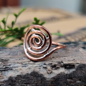 Può includere: Un anello color rame con un design a spirale. L'anello è appoggiato su un pezzo di legno. La spirale è il punto focale dell'anello, con una finitura liscia e lucida. La fascia dell'anello si curva per incontrare la spirale.