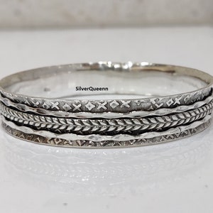 May include: Silver bangle bracelet with a woven design and multiple bands. The bracelet has a hammered finish and a textured surface.