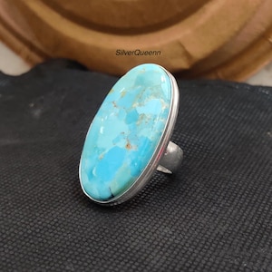 May include: A large oval turquoise ring with a silver band. The turquoise stone has a vibrant blue colour with brown and tan inclusions. The ring is set on a dark surface, with the text "SilverQueenn" visible.