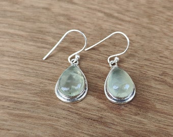 May include: A pair of silver teardrop earrings with pale green, translucent stones. The earrings have a simple, elegant design with a hook closure. The stones are set in a silver bezel.