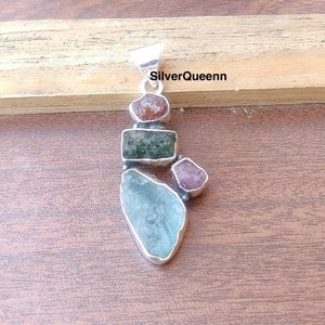 Pode incluir: Um pingente de prata com um grupo de pedras preciosas multicoloridas. O pingente é feito de prata e apresenta uma água-marinha azul claro, uma turmalina verde e duas turmalinas rosa. O pingente está colocado sobre uma superfície de madeira.