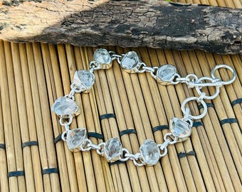 May include: A silver bracelet with clear, irregularly shaped gemstones. The bracelet features a toggle clasp and is displayed on a bamboo mat with a wooden accent and greenery in the background. The gemstones are set in silver bezels.