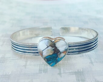 May include: A silver cuff bracelet with a heart-shaped turquoise and white stone inlay. The bracelet has three bands of silver with a rope-like design.