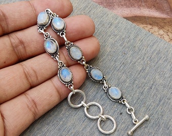 May include: A silver bracelet featuring oval moonstone cabochons. The bracelet has a toggle clasp and is composed of multiple links with bezel-set stones. The moonstones have a blue and white iridescent glow. The bracelet is displayed on a gray surface.