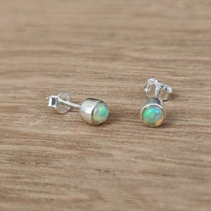 Puede incluir: Un par de pendientes de botón con engaste de plata. Cada pendiente presenta una piedra de ópalo redonda e iridiscente. Los pendientes se muestran sobre una superficie de madera marrón claro. Son pequeños y delicados.
