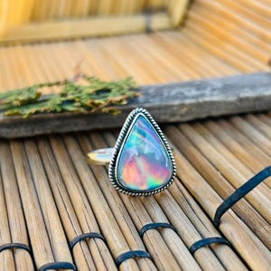May include: A silver ring with a pear-shaped, iridescent opal gemstone. The opal displays a range of colors, including blue, green, and pink. The ring is set on a wooden surface.