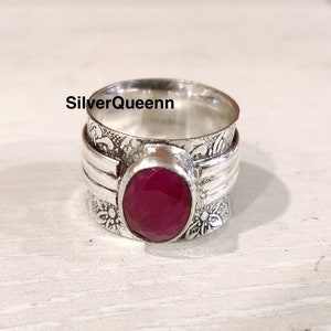 Puede incluir: Un anillo de plata con un diseño floral adornado y una gran piedra preciosa de rubí de forma ovalada. El anillo es de estilo spinner, con una banda que gira alrededor de la piedra preciosa.