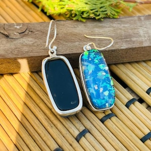 May include: A pair of silver earrings with rectangular, elongated pendants. One earring has a solid black stone, the other a vibrant blue and green mosaic design. The earrings are displayed on a wooden surface.