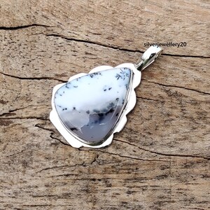 May include: A silver pendant with a white and gray dendritic agate stone. The stone is set in a silver bezel and has a scalloped edge. The pendant is on a wooden surface.