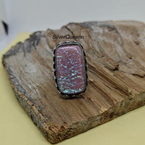Könnte beinhalten: Rechteckiger Ring mit einem rosa schillernden Stein, der in einen silberfarbenen Metallrahmen gefasst ist. Der Ring hat einen strukturierten Rand. Der Text "SilverQueenn" ist sichtbar.