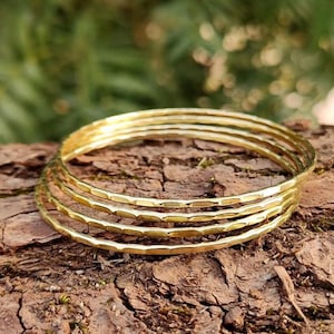 Puede incluir: Un conjunto de cuatro brazaletes circulares y delgados de tono dorado con una superficie martillada y texturizada. Las pulseras están apiladas, mostrando un acabado metálico reflectante con pequeñas hendiduras uniformes a lo largo de todo el borde.