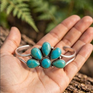 May include: A silver cuff bracelet featuring five teardrop-shaped turquoise stones arranged in a fan pattern. The central stone is round and surrounded by a twisted silver wire detail, while the four surrounding stones are set in smooth silver bezels.