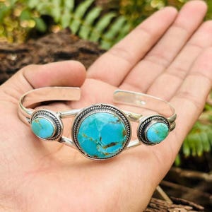 May include: A silver cuff bracelet featuring three round turquoise stones with brown matrix patterns. The central stone is the largest, flanked by two smaller stones, all set in twisted silver wire frames on a polished silver band.