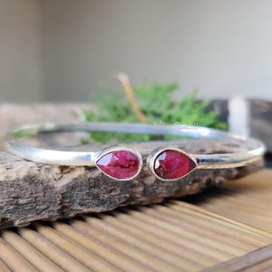 Puede incluir: Una pulsera de plata con dos gemas rojas en forma de lágrima. La pulsera tiene un diseño sencillo y elegante, ideal para el uso diario. Las gemas están engastadas en una montura de plata pulida.