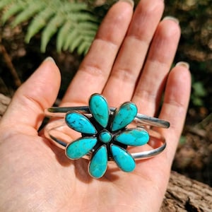 May include: A silver cuff bracelet featuring a central flower-shaped design composed of seven teardrop-shaped turquoise stones with dark matrix veining and a small round turquoise stone at the centre of the floral arrangement.
