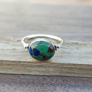 May include: A silver ring featuring an oval green, blue and brown gemstone. The ring has a textured band with small beads on the sides.