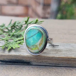 May include: A silver ring with an oval, iridescent, teal-colored stone. The stone has a unique pattern with hints of yellow and white. The ring is set on a wooden surface with green foliage in the background. The text "SilverQueenn" is visible.