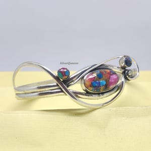 Op de afbeelding: Een zilveren manchetarmband met een abstract ontwerp. De armband heeft een ovaalvormige, veelkleurige steen met roze, blauwe en gouden tinten. Twee kleinere, ronde stenen zijn aan weerszijden van de centrale steen geplaatst. De tekst "SilverQueenn" is zichtbaar.