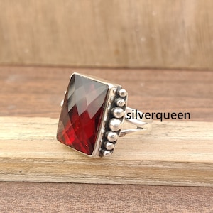 May include: A silver ring with a red gemstone set in a square bezel. The ring has a beaded band.