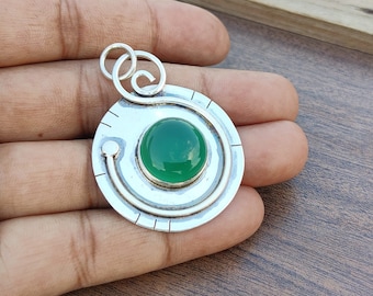 May include: A silver pendant with a green gemstone set in a circular design. The pendant has a swirl design and a loop for attaching a chain.