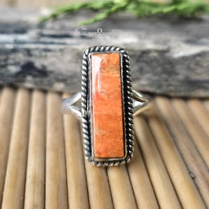 Può includere: Un anello d'argento con una pietra arancione rettangolare. La pietra è incastonata in una cornice d'argento con un motivo a corda intrecciata. L'anello è su una superficie di legno.