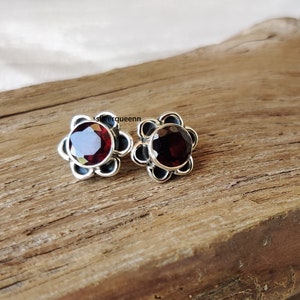 Op de afbeelding: Een paar zilveren oorbel studs met rode edelsteen accenten. Elke oorbel heeft een ronde edelsteen die is gezet in een bloemvormig ontwerp.