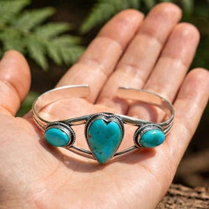 May include: A silver cuff bracelet featuring a central heart-shaped turquoise stone flanked by two smaller oval turquoise stones. The metal band has a polished finish, and each stone is set within a decorative twisted rope-style silver border.