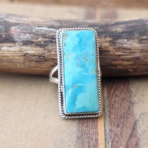 May include: A rectangular ring featuring a bright blue turquoise stone with natural brown matrix patterns. The stone is set in a polished silver frame with a twisted rope border design and a split shank band that tapers toward the back of the ring.