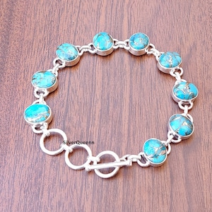 May include: A silver bracelet with oval turquoise stones, each with a gold-coloured matrix pattern. The bracelet has a toggle clasp and is displayed on a brown wooden surface. The brand name "SilverQueenn" is visible.