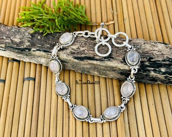 Op de afbeelding: Een zilveren armband met ovale rozenkwarts stenen. De stenen zijn gezet in een zilveren bezel met een gedraaid touwontwerp. De armband heeft een tuimelsluiting en wordt gepresenteerd op een houten oppervlak.