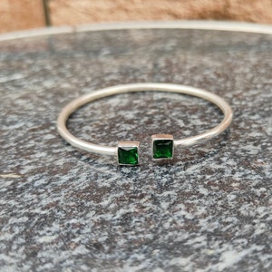 Op de afbeelding: Een zilveren armband met twee vierkante groene edelstenen.