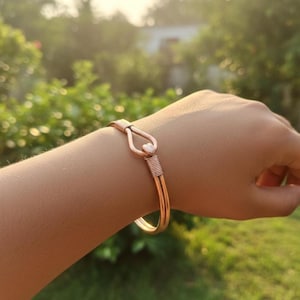 Puede incluir: Una pulsera de color cobre con un cierre en forma de corazón. La pulsera presenta un diseño de alambre enrollado y se lleva en la muñeca. El fondo es una vista borrosa de follaje verde.