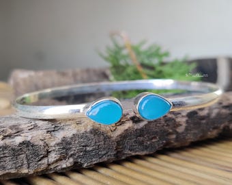May include: A silver-toned cuff bracelet with two teardrop-shaped, bright turquoise-colored stones. The bracelet is displayed on a piece of weathered wood, with a blurred background of greenery.