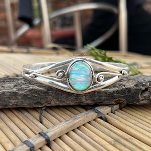 Puede incluir: Un brazalete de plata con una piedra de ópalo ovalada e iridiscente en el centro. El brazalete tiene un diseño delicado con detalles en espiral y se muestra sobre un trozo de madera.