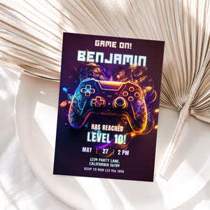 Puede incluir: Un mando de videojuego morado y azul con luces de neón, una invitación de cumpleaños para el décimo cumpleaños de Benjamin. La invitación dice "Game On! Benjamin Has Reached Level 10!" con la fecha y la hora de la fiesta.