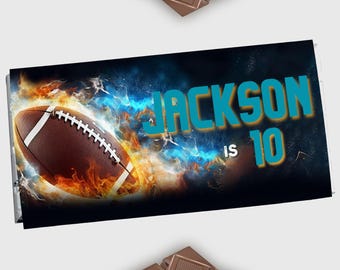 May include: A personalized candy bar wrapper with the name "Jackson" and the number "10" in teal. A brown football is set against a fiery orange and blue background. The wrapper is surrounded by chocolate bars.