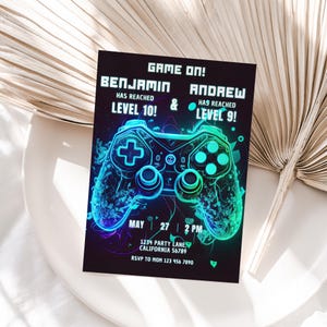 May include: A black and blue video game party invitation with a neon green and blue video game controller graphic. The text reads "Game On! Benjamin has reached Level 10! & Andrew has reached Level 9!" with the date and time of the party, May 27, 2 PM, and the address 1234 Party Lane, California 56789. The invitation also includes the RSVP information, "RSVP to Mom 123 456 7890".