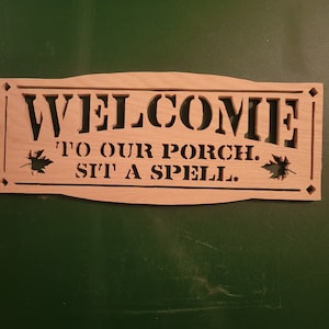 May include: Wooden sign with the text "WELCOME TO OUR PORCH. SIT A SPELL." in a decorative font. The sign is painted in a light brown color and has a distressed look. There are two leaf designs on either side of the text.