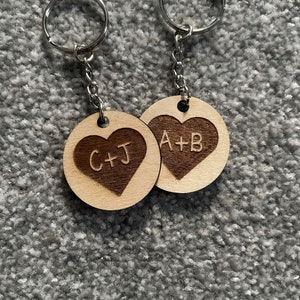 Könnte beinhalten: Zwei Schlüsselanhänger aus Holz in Herzform mit gravierten Initialen. Der Schlüsselanhänger links sagt "C+J" und der Schlüsselanhänger rechts sagt "A+B".