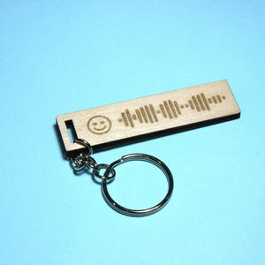 Peut inclure: Un porte-cl&eacute;s en bois avec un visage souriant et un motif d'onde sonore. Le porte-cl&eacute;s est attach&eacute; &agrave; un anneau de cl&eacute; argent&eacute;.