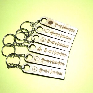 Könnte beinhalten: Ein Satz von sieben Holzschlüsselanhängern mit verschiedenen Symbolen und einem Barcode-ähnlichen Design. Die Schlüsselanhänger sind an silbernen Schlüsselringen befestigt.
