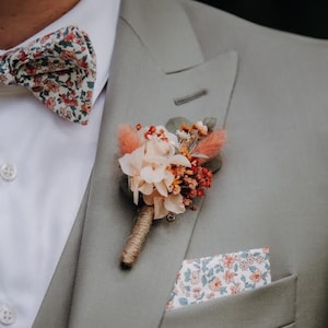 Op de afbeelding: Een grijs colbert met een strik met bloemenpatroon, een zakdoek met bloemenpatroon en een klein boeket gedroogde bloemen die op de revers is geprikt.