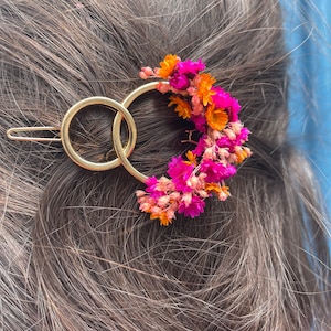 Pode incluir: Um grampo de cabelo de metal dourado com dois círculos entrelaçados, decorado com flores secas rosa e laranja.