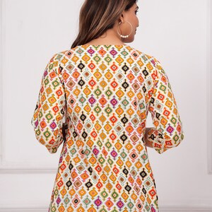 K&ouml;nnte beinhalten: Ein gemustertes Tunika-Oberteil mit einem sich wiederholenden geometrischen Design in Orange, Gelb, Gr&uuml;n, Lila und Schwarz auf wei&szlig;em Hintergrund. Das Oberteil hat Dreiviertel&auml;rmel und einen V-Ausschnitt. Das Model tr&auml;gt eine blaue Jeans.