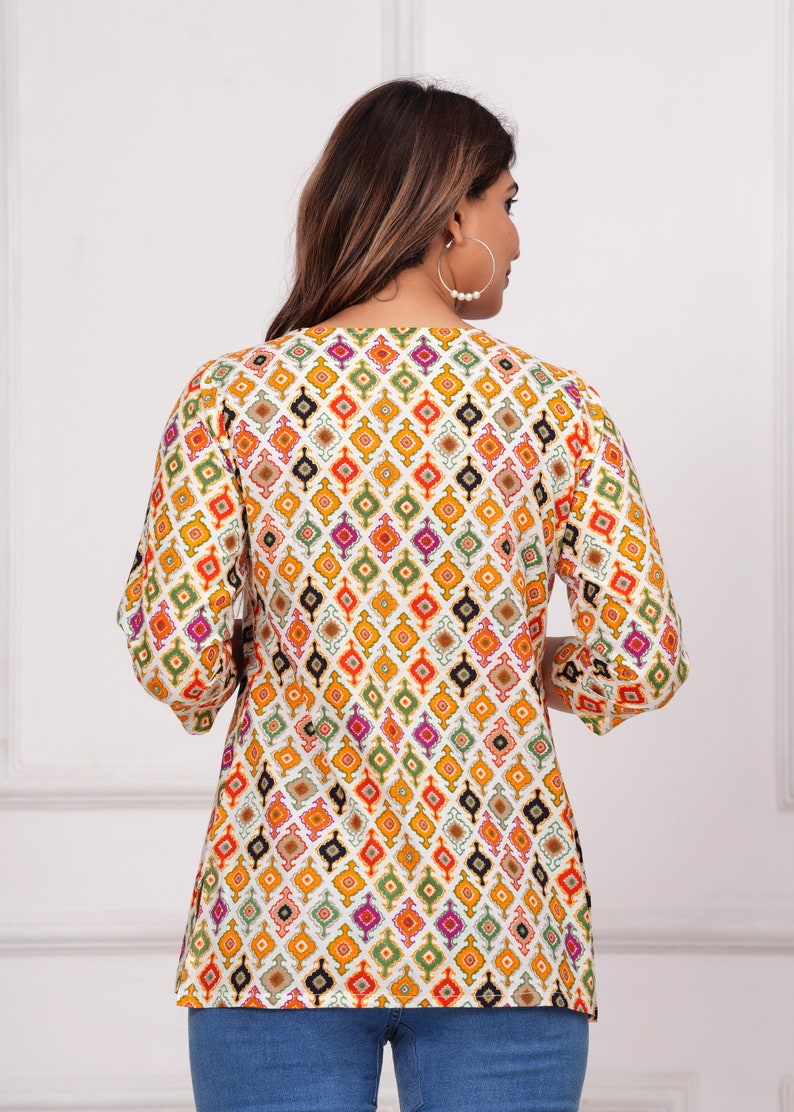 K&ouml;nnte beinhalten: Ein gemustertes Tunika-Oberteil mit einem sich wiederholenden geometrischen Design in Orange, Gelb, Gr&uuml;n, Lila und Schwarz auf wei&szlig;em Hintergrund. Das Oberteil hat Dreiviertel&auml;rmel und einen V-Ausschnitt. Das Model tr&auml;gt eine blaue Jeans.