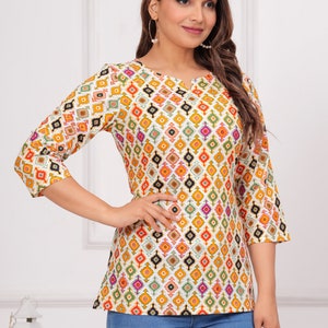 K&ouml;nnte beinhalten: Ein wei&szlig;es Oberteil mit einem farbenfrohen geometrischen Muster in Orange, Gelb, Gr&uuml;n und Rosa. Das Oberteil hat Dreiviertel&auml;rmel und einen Rundhalsausschnitt. Die Person tr&auml;gt eine blaue Jeans.