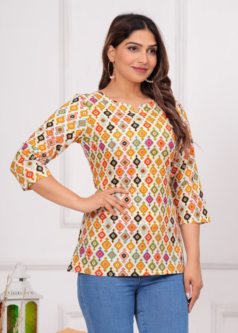 K&ouml;nnte beinhalten: Ein wei&szlig;es Oberteil mit einem farbenfrohen geometrischen Muster in Orange, Gelb, Gr&uuml;n und Rosa. Das Oberteil hat Dreiviertel&auml;rmel und einen Rundhalsausschnitt. Die Person tr&auml;gt eine blaue Jeans.
